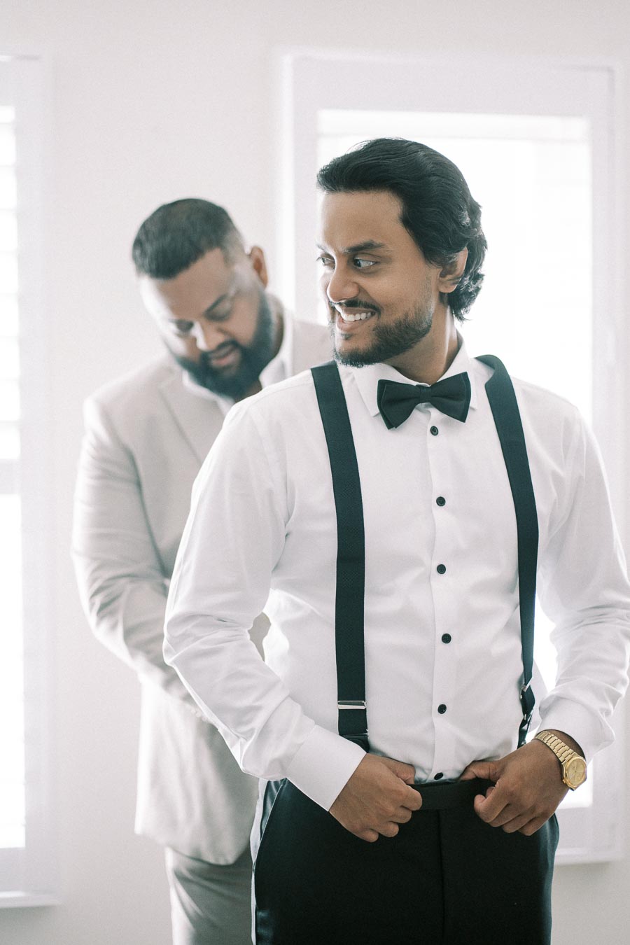 Two men preparing for a formal event, one adjusting his suspenders in a white shirt and black bow tie, the other in a light suit jacket assisting him in a brightly lit room.