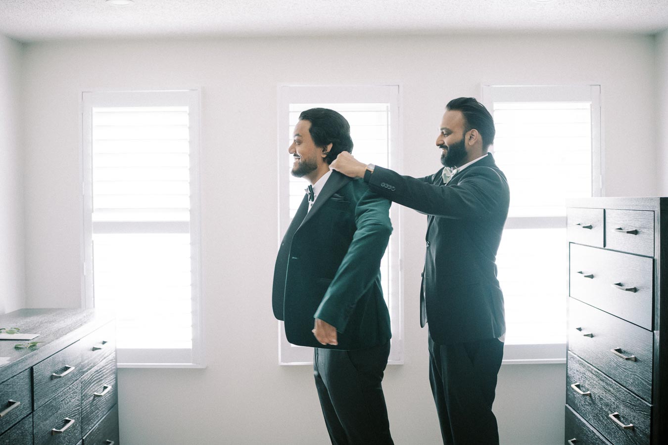 Two men dressed in formal suits prepare for a special occasion in a bright room with large windows. One man assists the other with his jacket, expressing camaraderie and attention to detail, as they stand near dark wooden dressers.