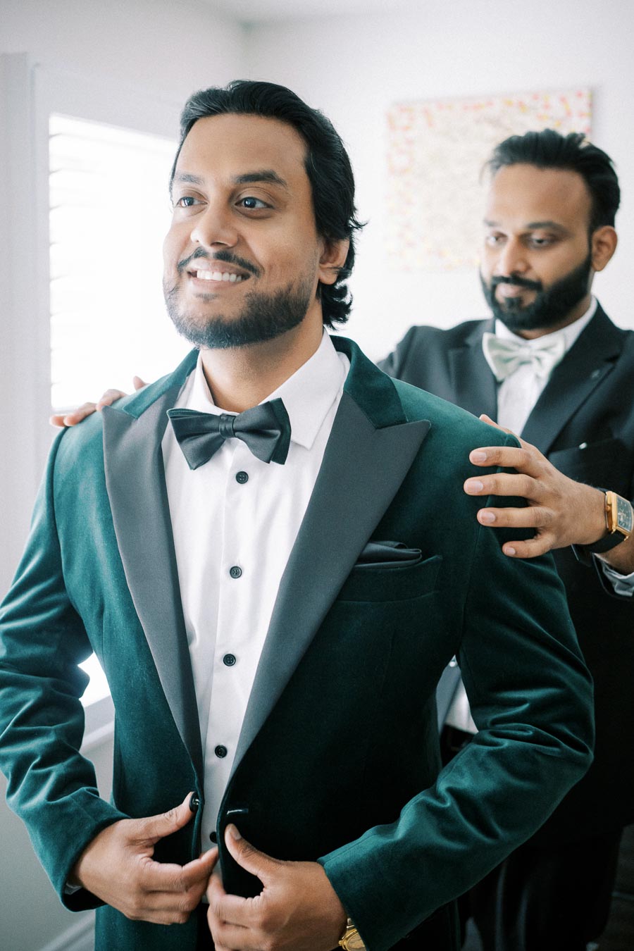 Groom in elegant green suit with black lapel smiling as a friend adjusts his bow tie, preparing for a wedding ceremony.