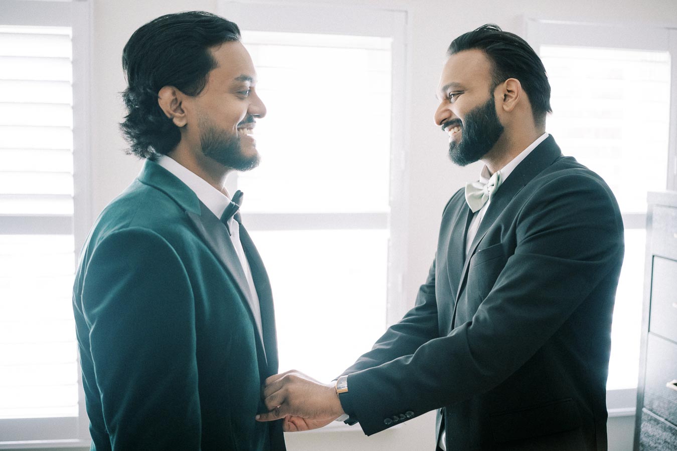 Two men in formal suits smiling at each other, standing in front of a bright window. One is adjusting the other's jacket, creating a warm and friendly atmosphere.