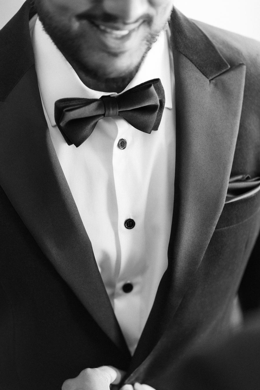 Black and white image of a man in a sharp tuxedo with a bow tie, smiling as he adjusts his jacket, highlighting elegant wedding or formal event attire.