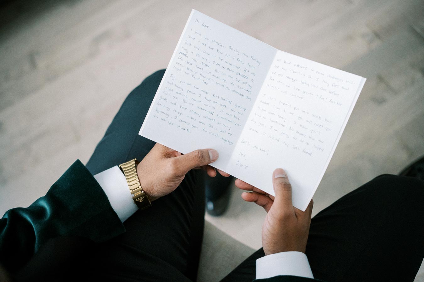 Man in a suit holding an open handwritten letter, seated on a wooden floor, wearing a gold watch.