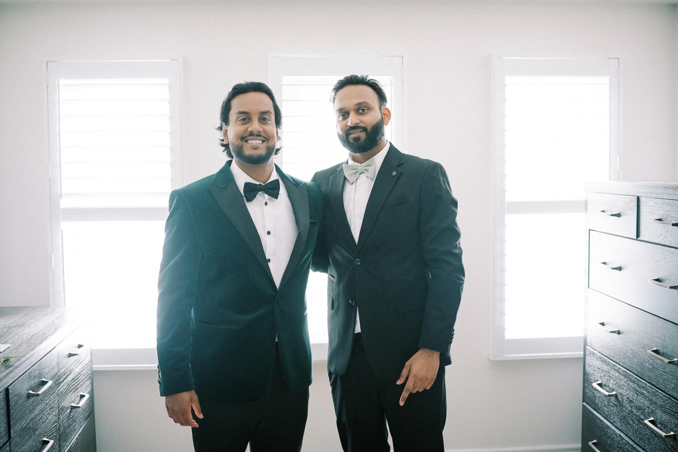 Two men in formal black suits and bow ties standing in a well-lit room with dressers and large windows.