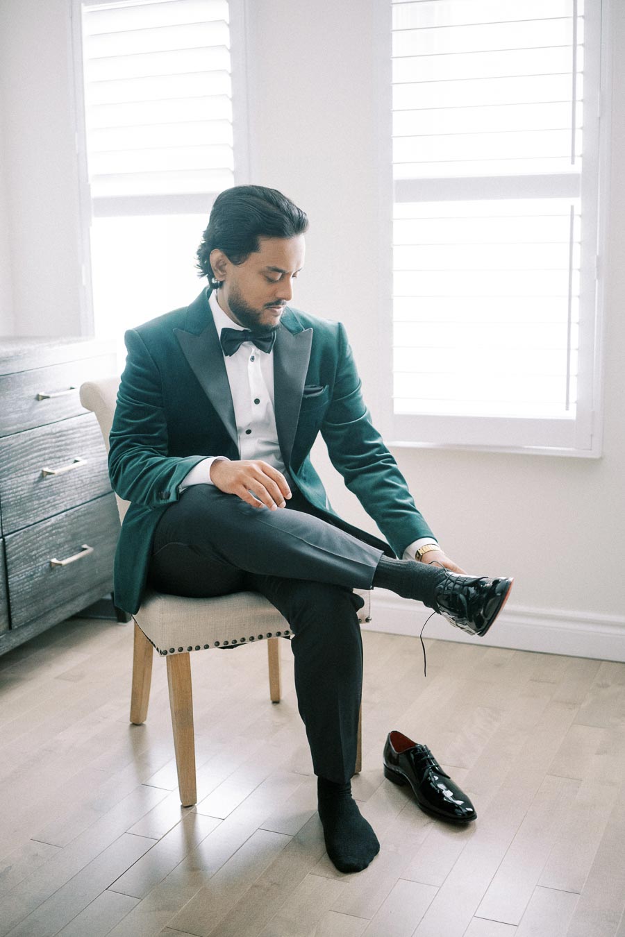 Man in a green tuxedo sitting on a chair, adjusting his black dress shoe, with natural light flowing through window shutters in a modern room.