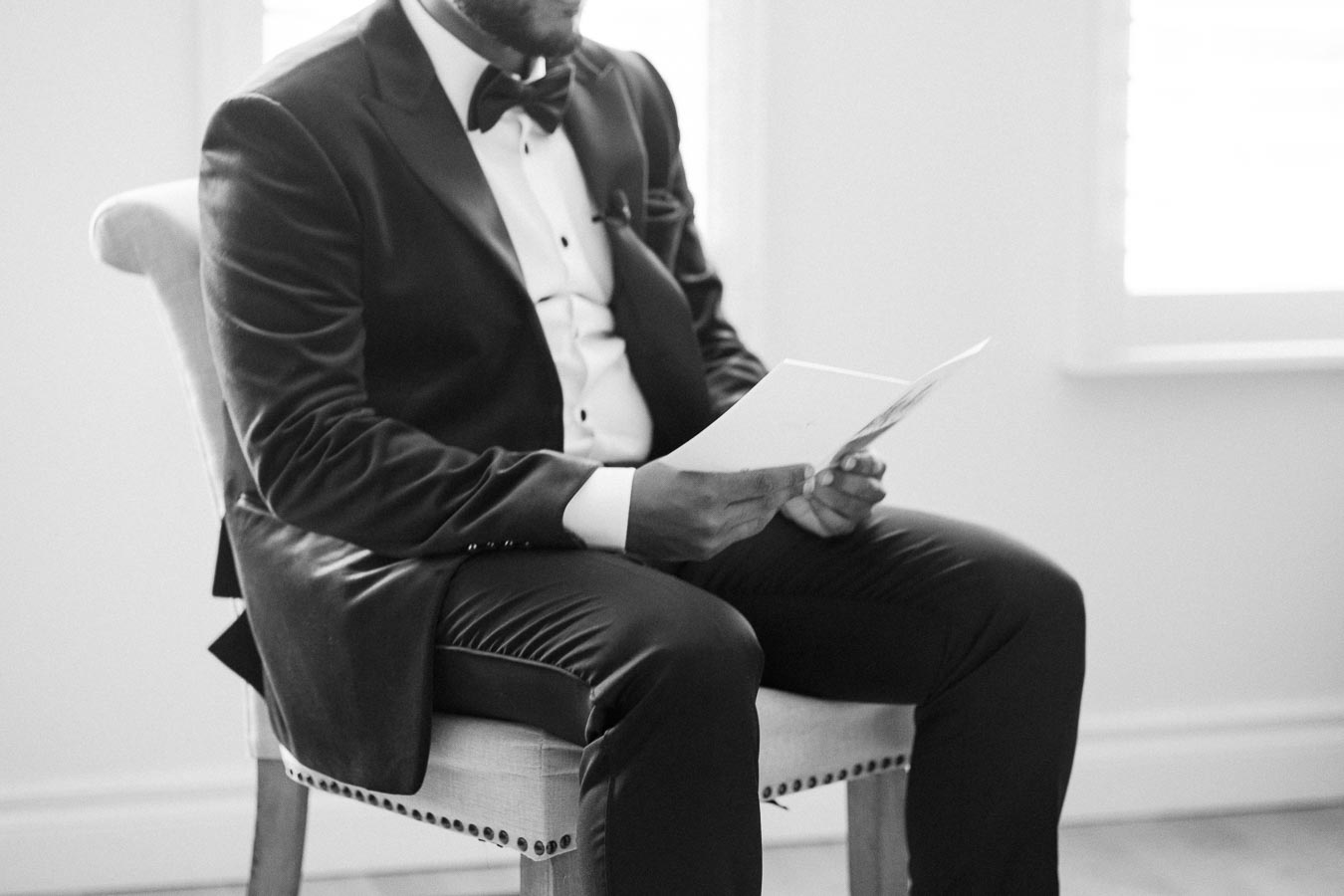 A man in a suit and bow tie sits on a chair while reading a card or letter, captured in a black-and-white setting.