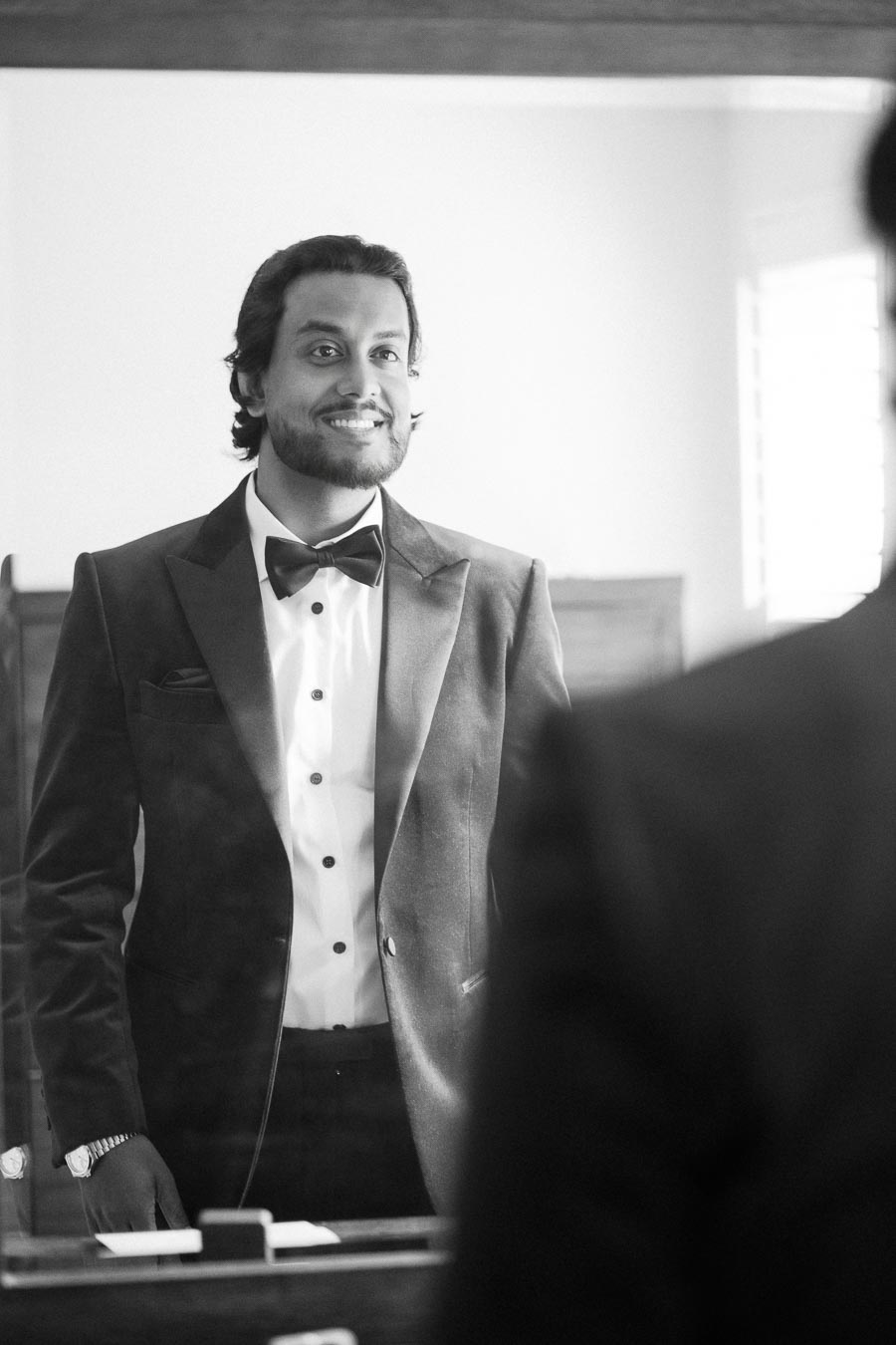 A man smiling while looking at himself in the mirror, wearing a formal black tuxedo with a bow tie.