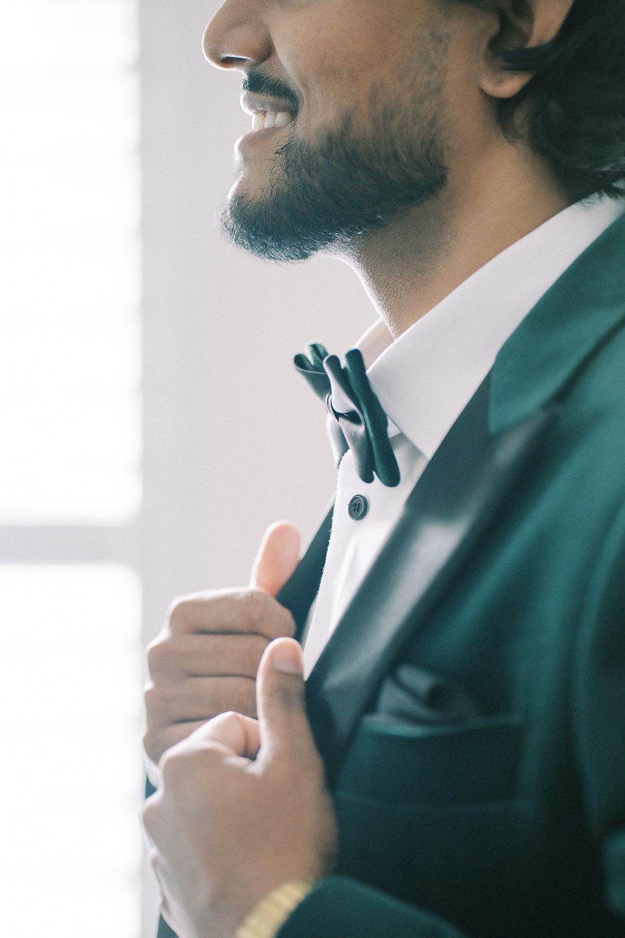 A man adjusting his bow tie, dressed in a formal black suit, smiling confidently.