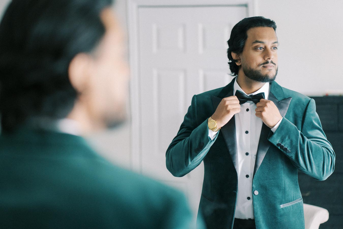 A man in a green velvet tuxedo adjusting his bow tie while looking in the mirror, preparing for a formal event.