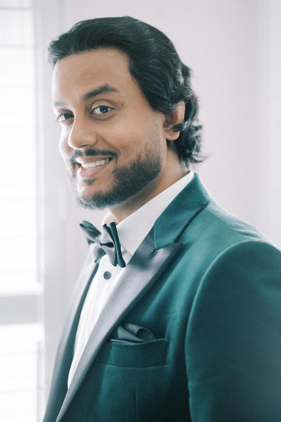 A man in a tuxedo smiling confidently, wearing a black bow tie and a dark green jacket, standing in front of a softly lit background.
