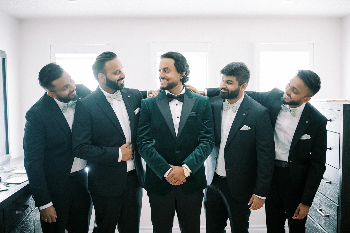 A group of five groomsmen wearing black suits and bow ties, smiling and standing in a well-lit room, celebrating a wedding day.