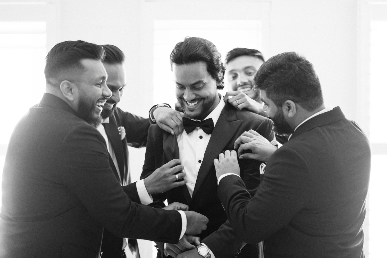 A groom surrounded by his groomsmen, all dressed in suits, laughing and preparing for a wedding ceremony.
