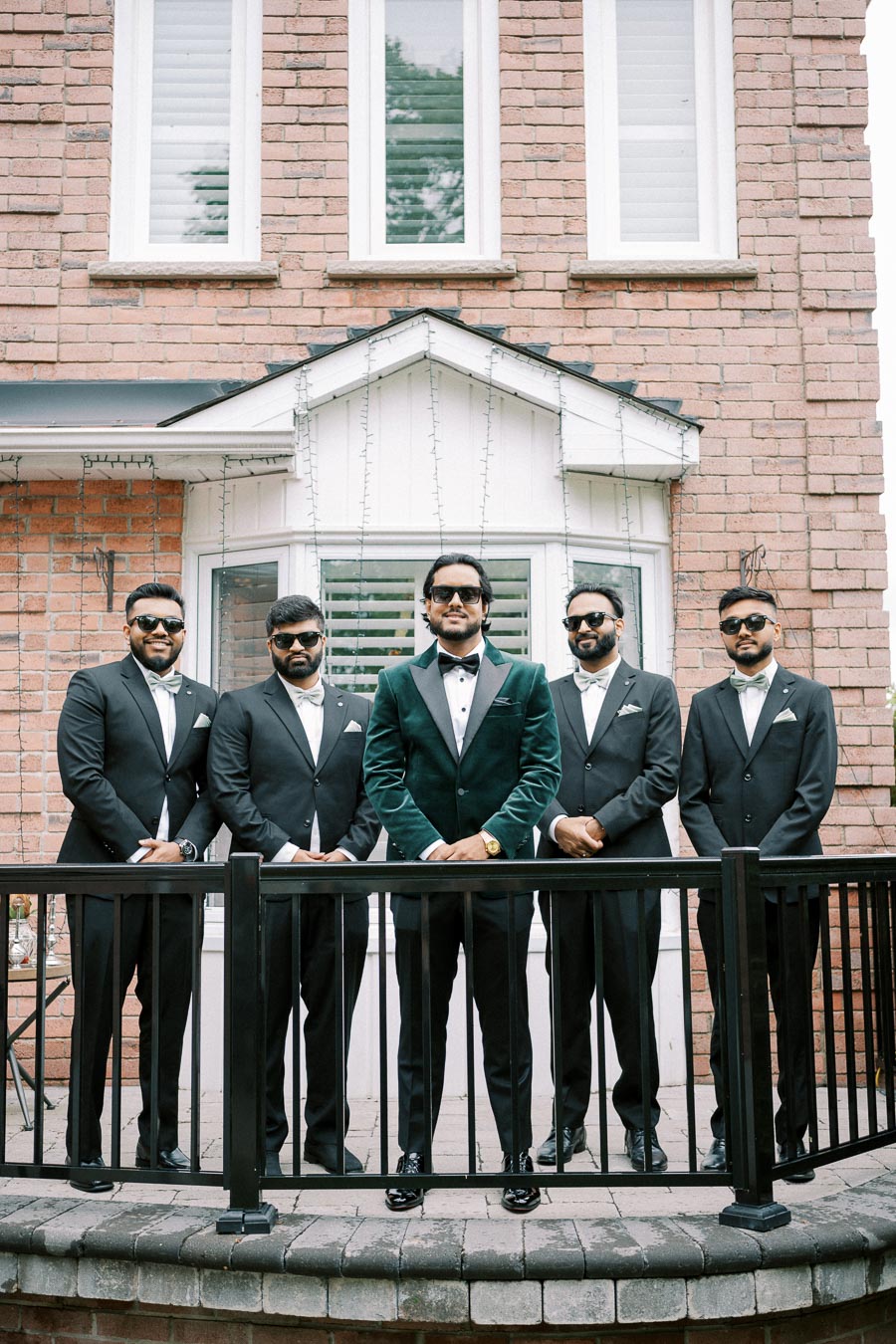 A group of five groomsmen in formal attire, wearing sunglasses, poses confidently on a brick patio in front of a brick house with white-framed windows.