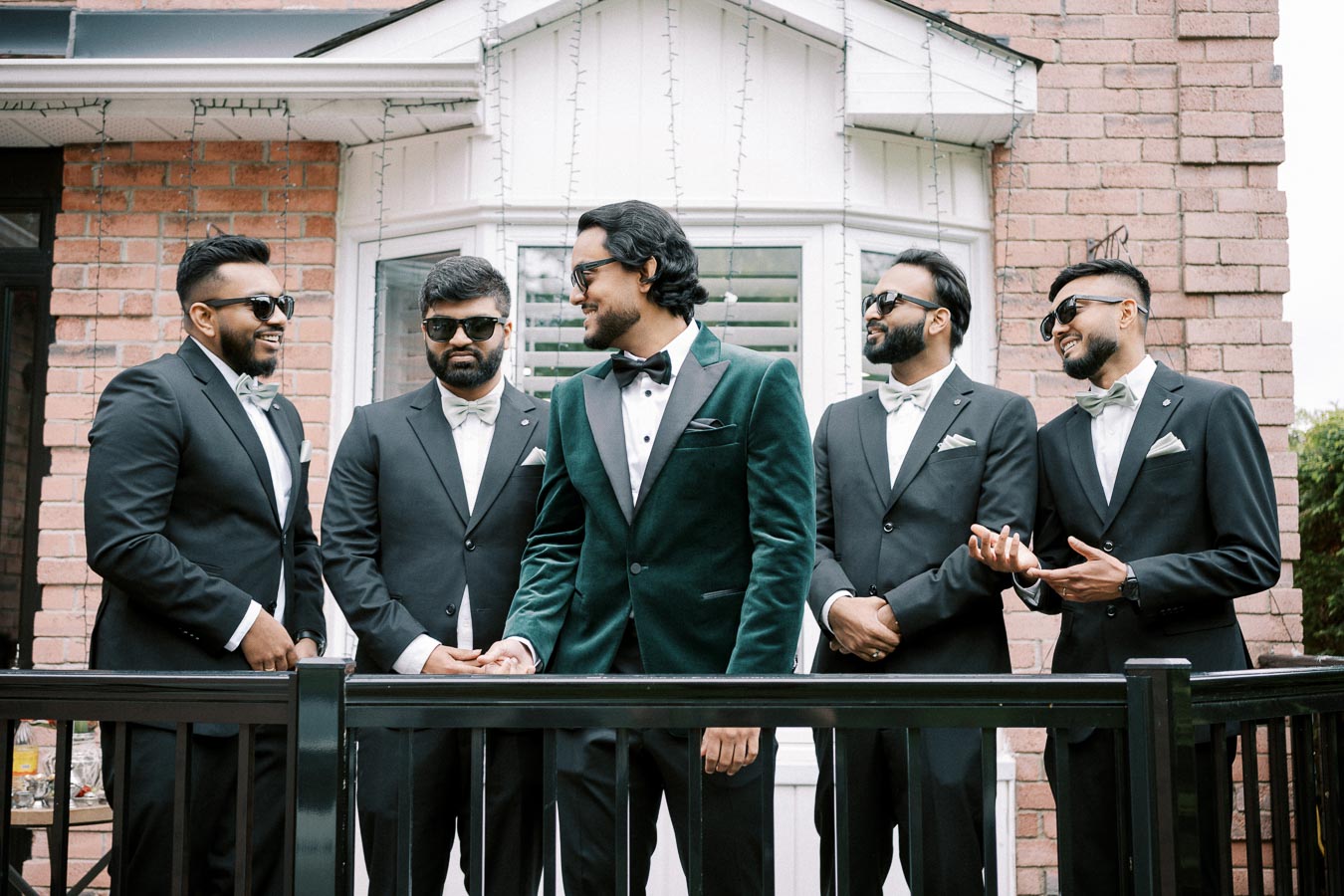 A group of five men in formal suits and sunglasses gather and smile outside a building. One man is wearing a green velvet tuxedo, standing out among the others dressed in black suits. The scene conveys a stylish and celebratory atmosphere, possibly a wedding or formal event.