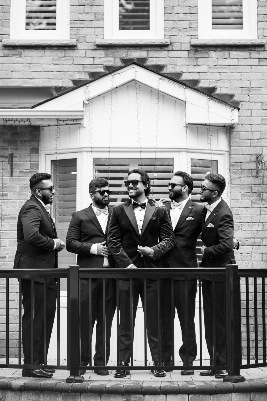 A group of five groomsmen in matching black suits pose together, smiling and wearing sunglasses, in front of a brick house with decorative lights.