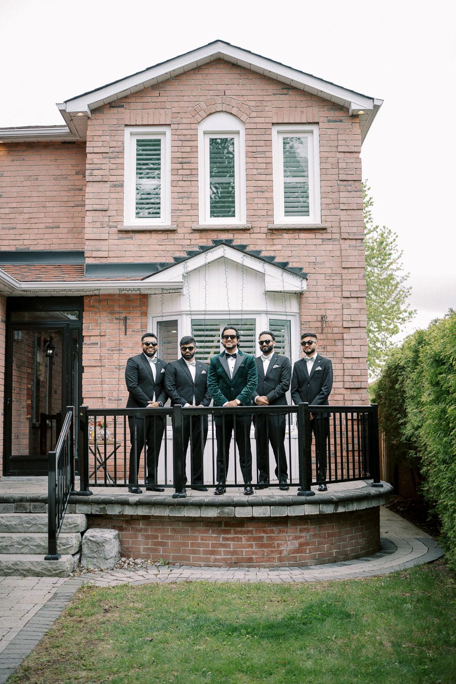 A group of five groomsmen wearing black suits and sunglasses stand on a porch in front of a brick house, exuding a formal and stylish vibe, perfect for a wedding or special event.