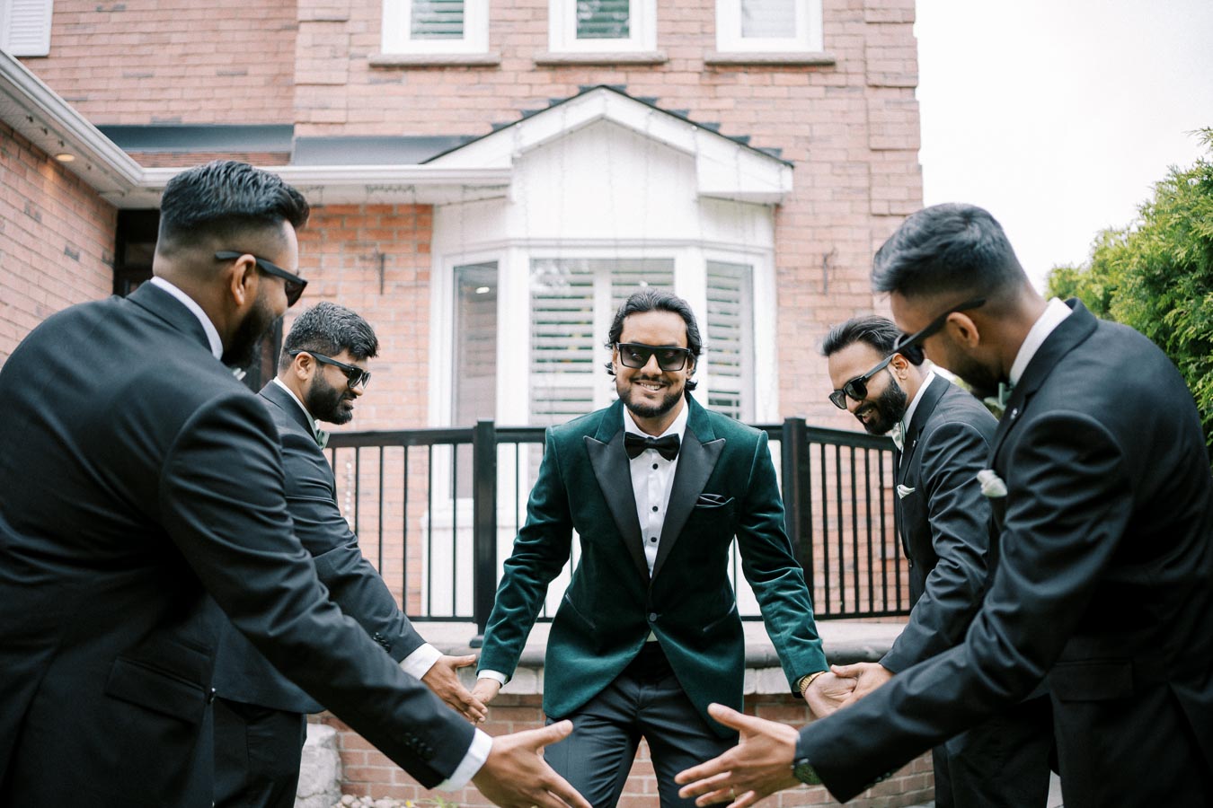 A group of five groomsmen in formal suits, smiling and wearing sunglasses, gathered in front of a brick house, preparing for a wedding ceremony.