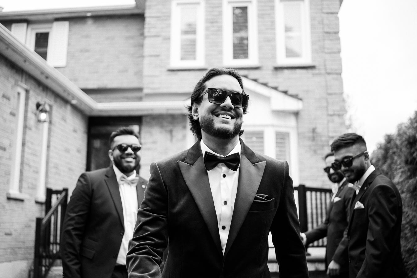 Black and white photo of a group of men dressed in formal suits and sunglasses, smiling and walking outside a brick building, conveying a stylish and confident mood.