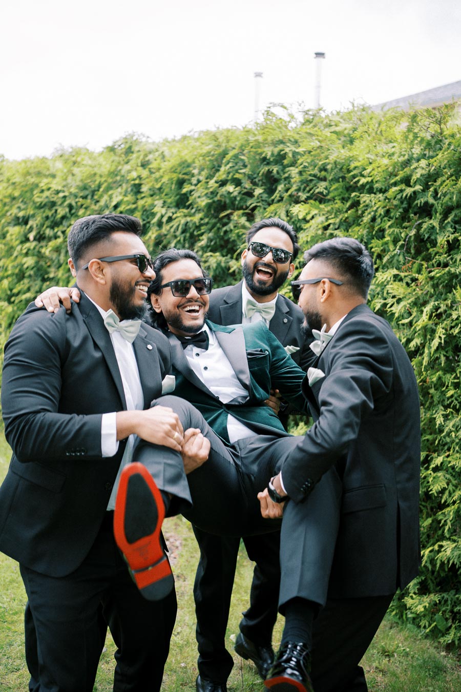 Group of four men in suits laughing and having fun outdoors. Three of them are holding up their friend, who is wearing sunglasses and a suit with a green jacket, in a playful manner in front of lush green foliage.