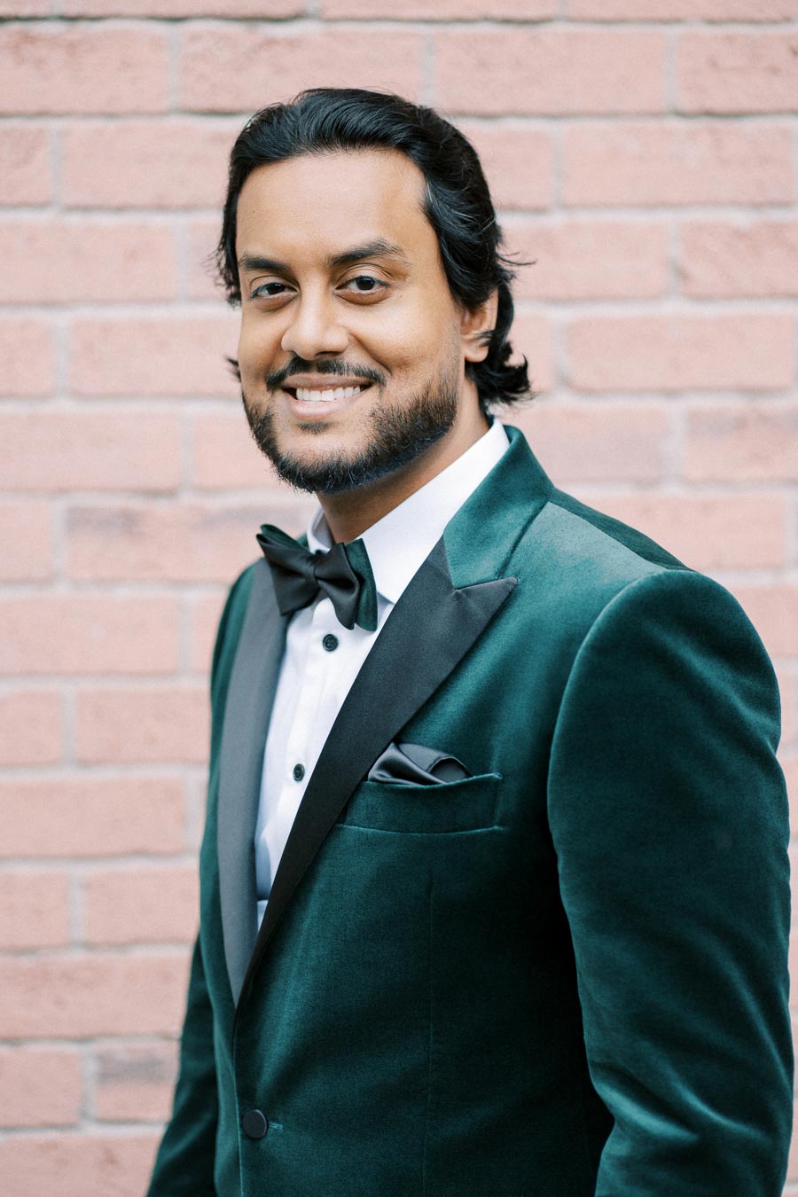 A man in a stylish green velvet suit and black bow tie smiling against a brick wall background.