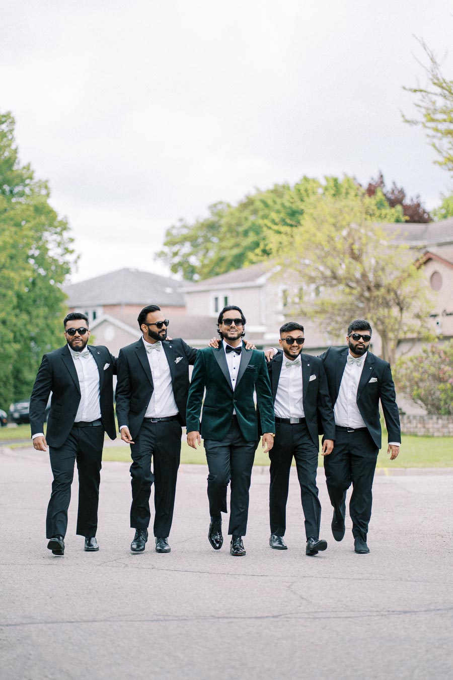 A group of five men in formal suits walking outdoors, smiling and appearing confident, with trees and houses in the background, capturing a stylish and cheerful moment.