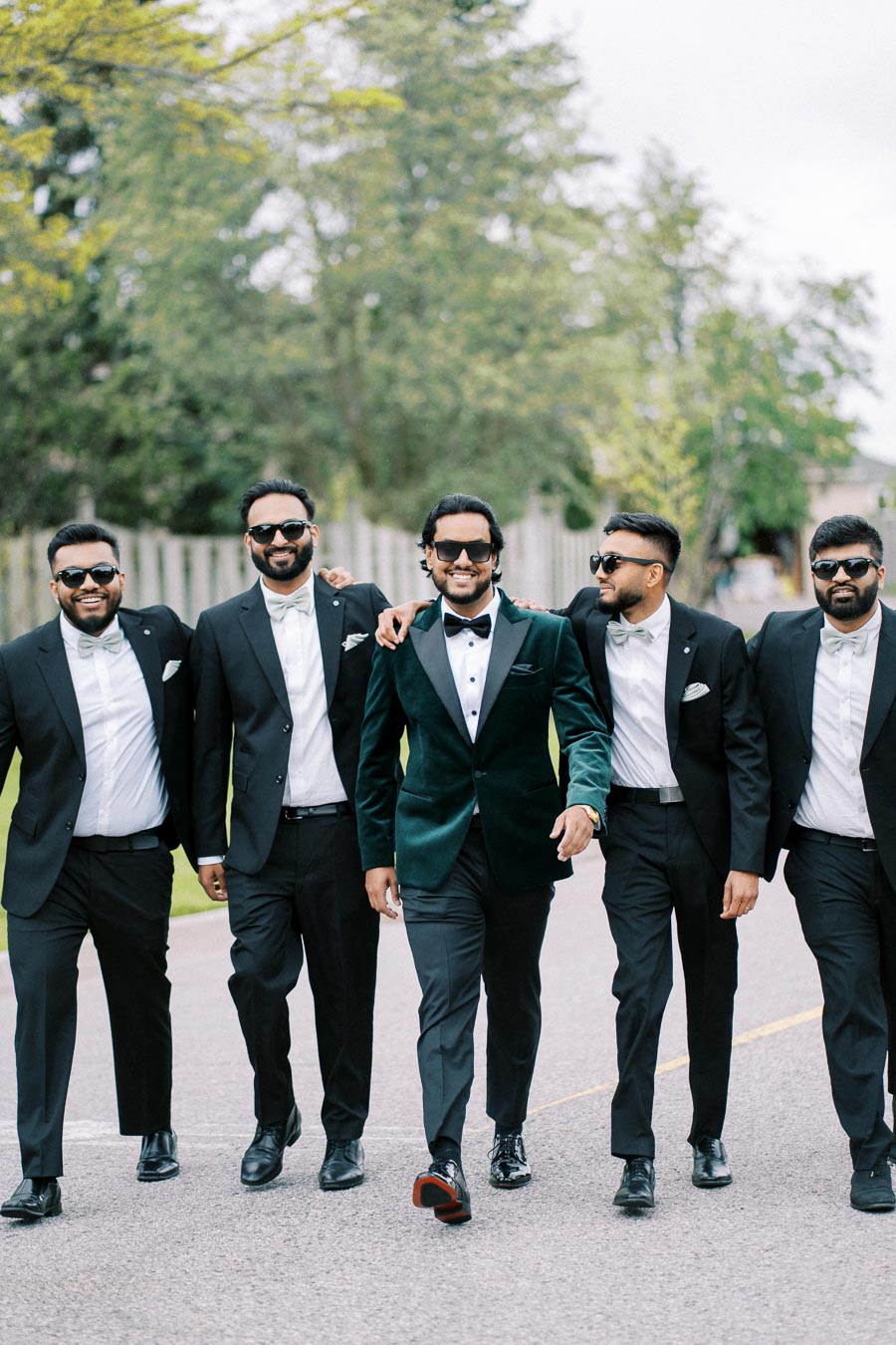 Five men in formal suits, wearing sunglasses and smiling, walking outdoors on a sunny day with trees in the background.