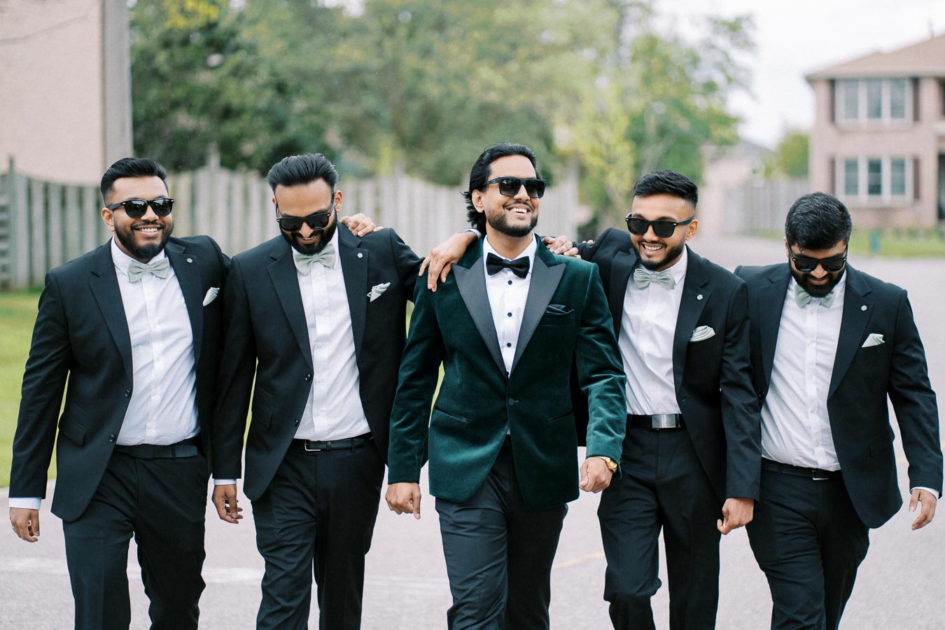Five men wearing tuxedos and sunglasses walking confidently outdoors, smiling and embracing each other, showcasing friendship and celebration.