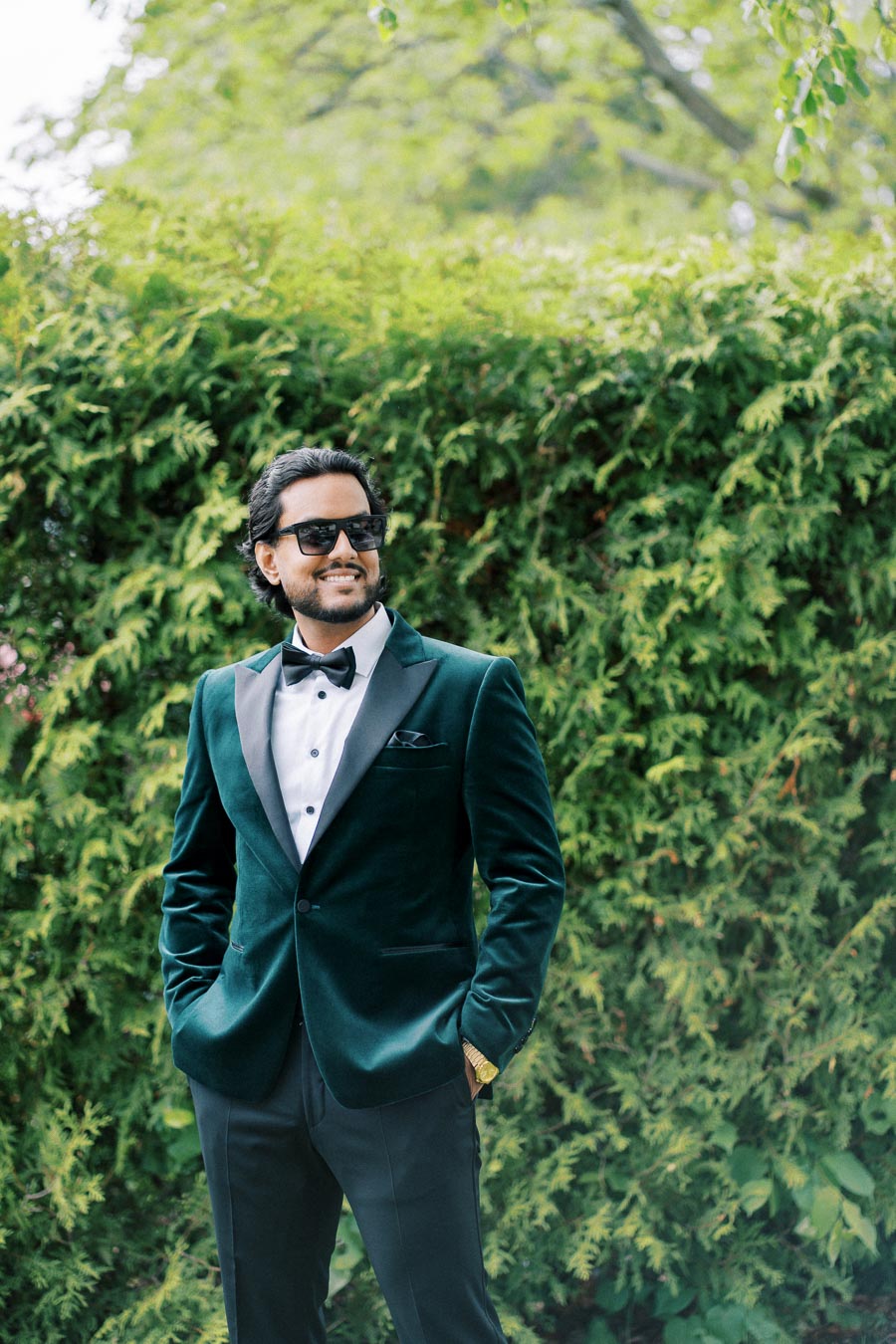 A stylish man in a dark green velvet suit and sunglasses, smiling confidently while standing in front of lush greenery.