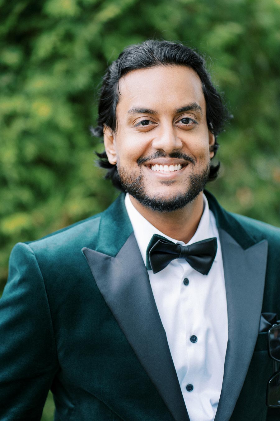 A man in a green tuxedo and black bow tie smiling in front of a lush green background.
