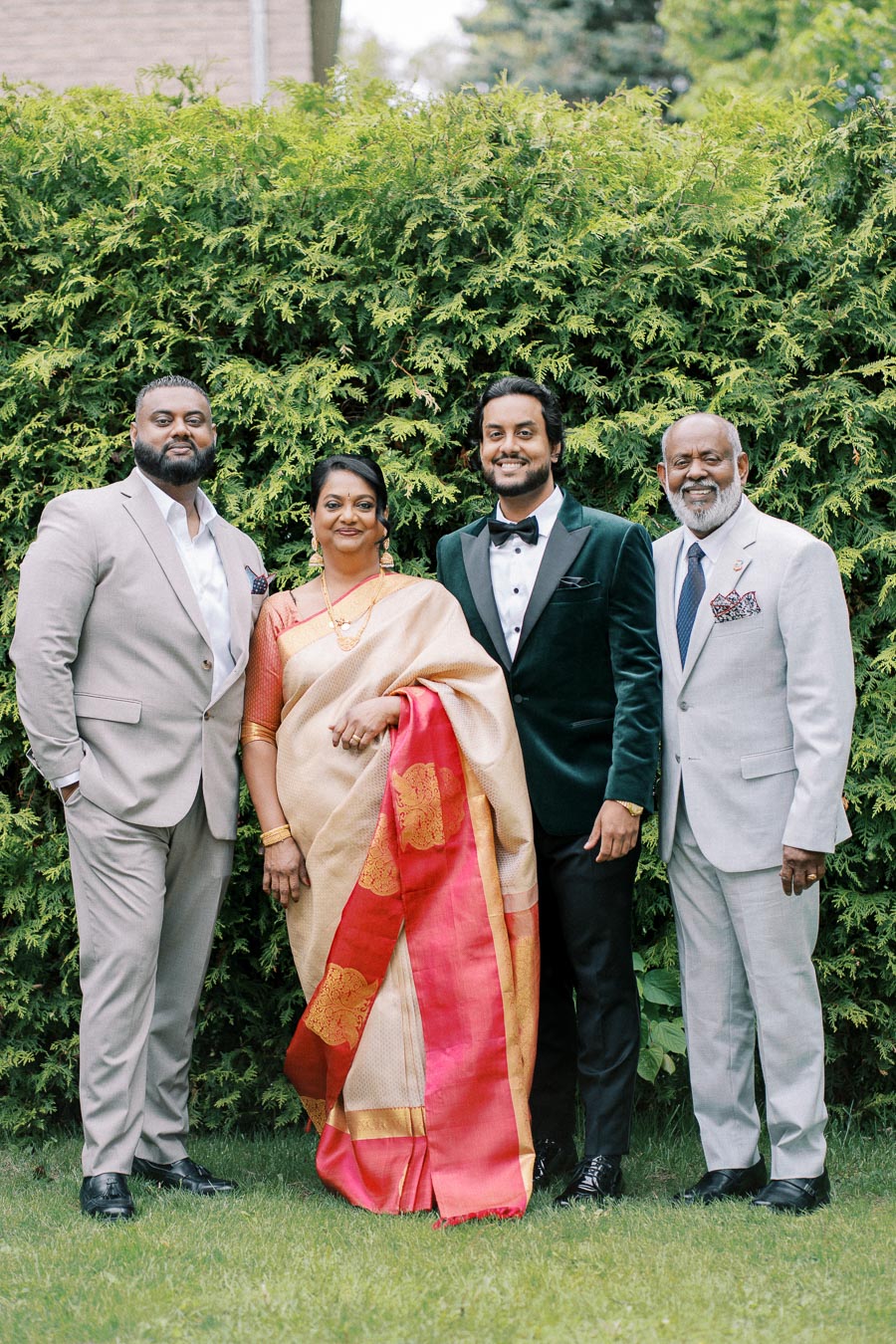 A family poses elegantly in formal attire, with two men in suits and a woman in a traditional saree, standing against a lush green hedge backdrop.