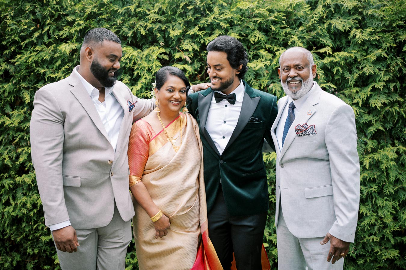 A family of four, dressed in formal attire, stands together in front of lush greenery. The woman is wearing a traditional sari, while the men are in suits, creating a harmonious and joyful group portrait.