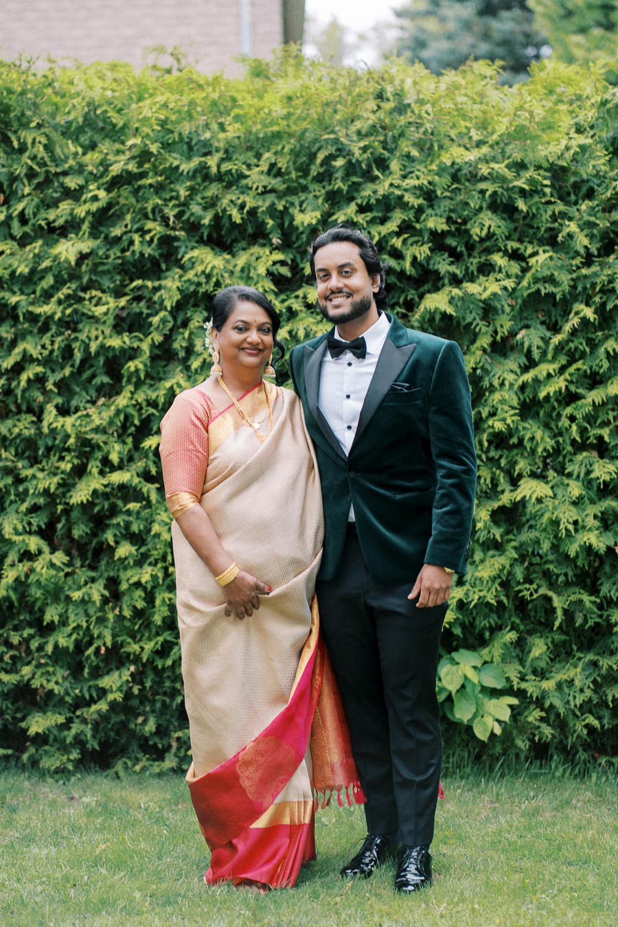 A smiling couple poses outdoors, with the woman wearing a traditional saree and the man in a formal green velvet suit, standing in front of a lush green hedge.