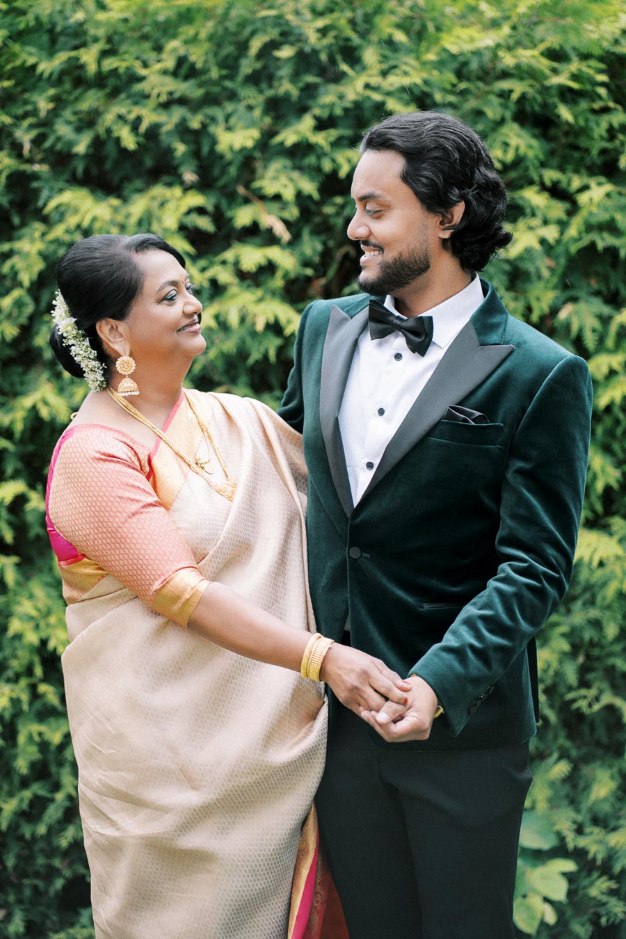 Smiling man in a green velvet tuxedo holding hands with a woman in a traditional beige and pink sari, standing in front of lush greenery.