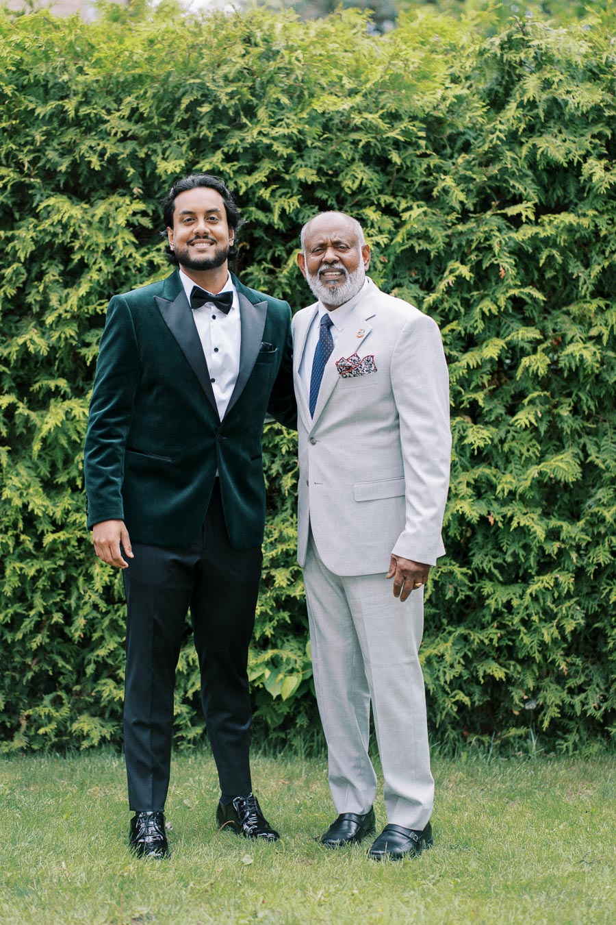 Two elegantly dressed men posing in formal suits with green foliage background.
