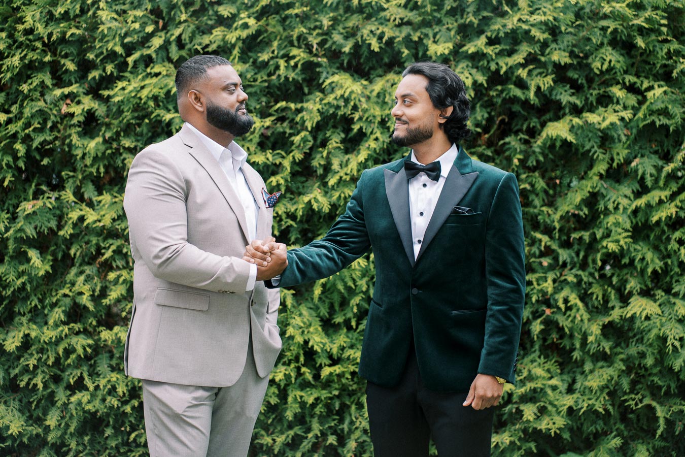 Two men in formal attire shaking hands in front of a green foliage background, showcasing a gesture of friendship and agreement. One is wearing a light beige suit, while the other is in a dark velvet tuxedo with a bow tie.
