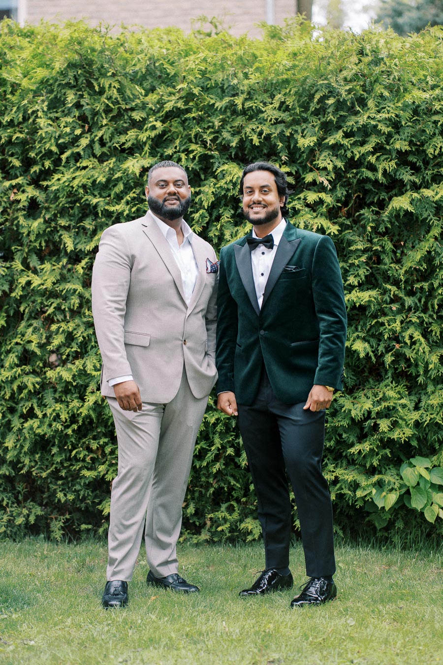 Two men in formal attire, one in a beige suit and the other in a dark green velvet tuxedo, smiling and standing on grass with a lush green hedge background.