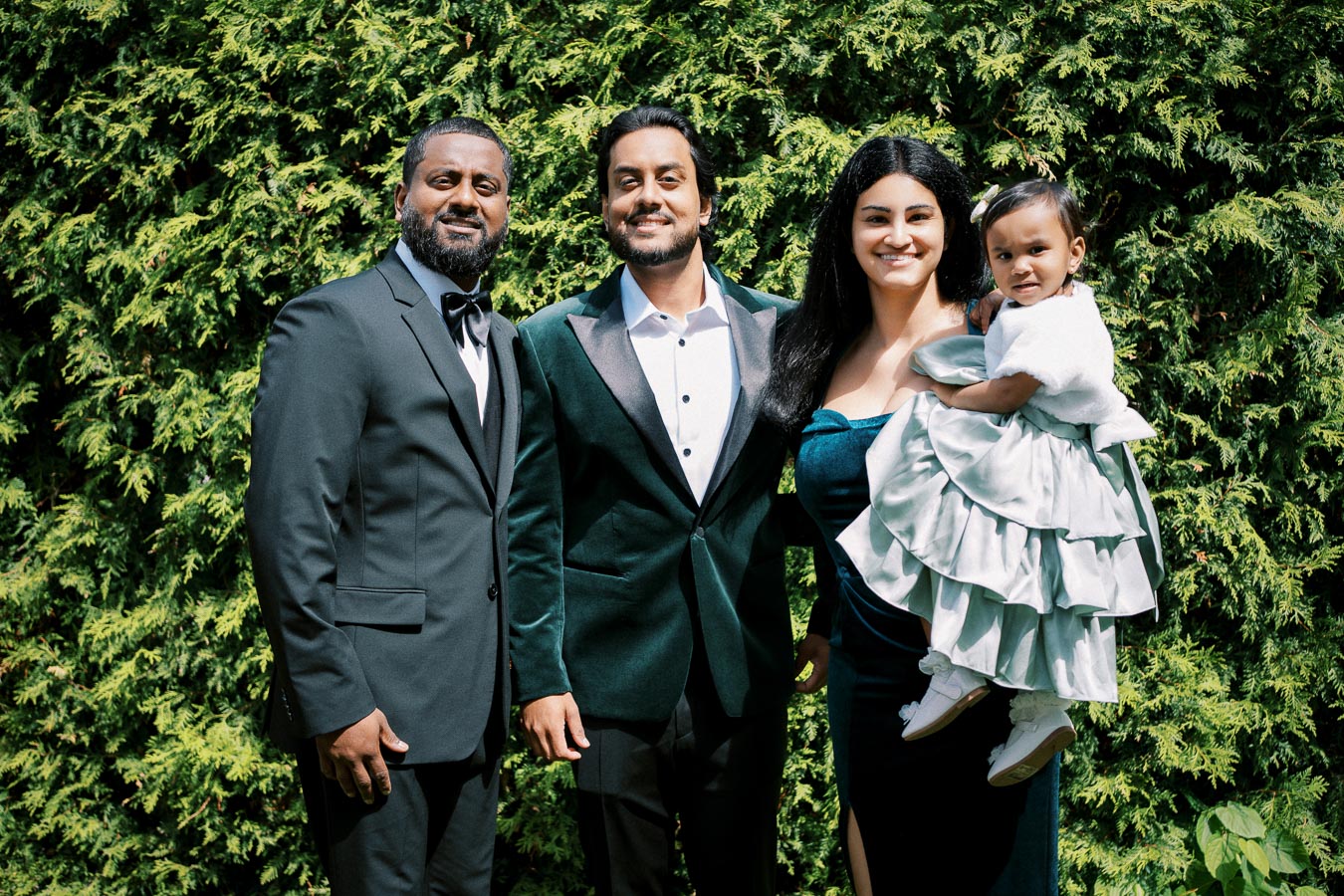 A group of four people, including a child, dressed formally and smiling in front of lush green foliage.