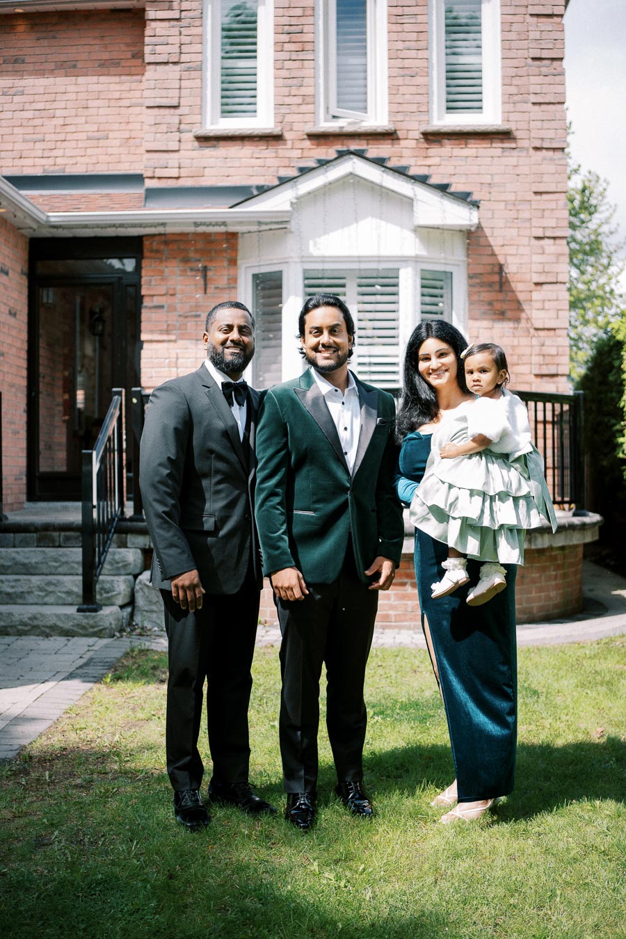 A family standing in front of a brick house on a sunny day, with two men in suits, a woman in a blue dress holding a baby girl in a green dress.