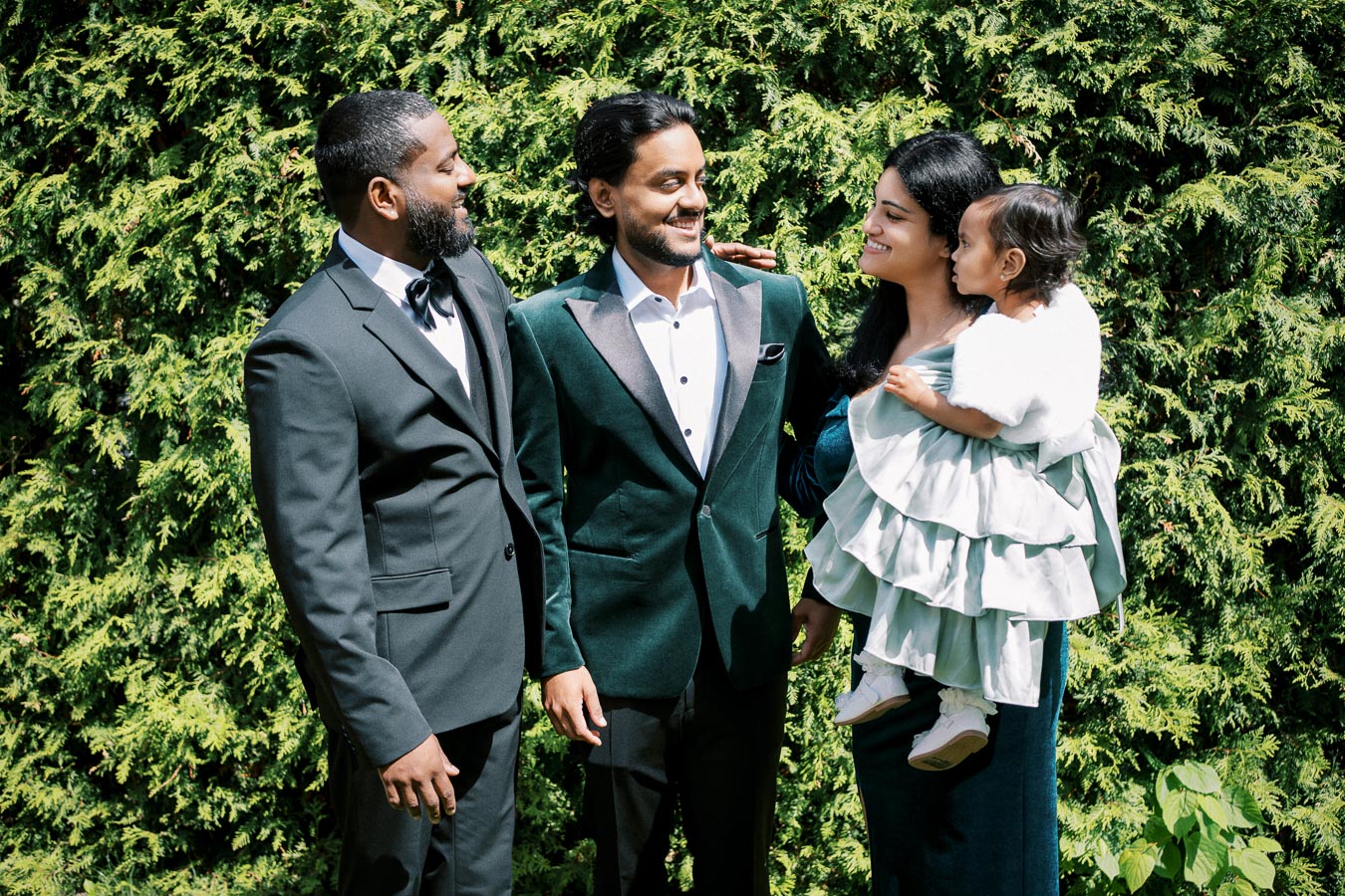 A joyful group of four people dressed in formal attire, standing together outdoors in front of lush greenery, with a woman holding a young child wearing a ruffled dress.