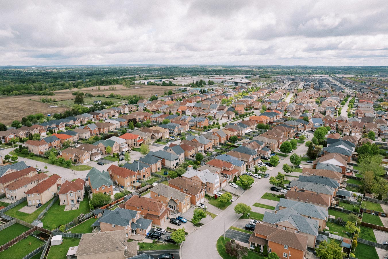 Aerial view of a suburban neighborhood showcasing rows of houses with varied roof colors, surrounded by green lawns and trees, under a cloudy sky.