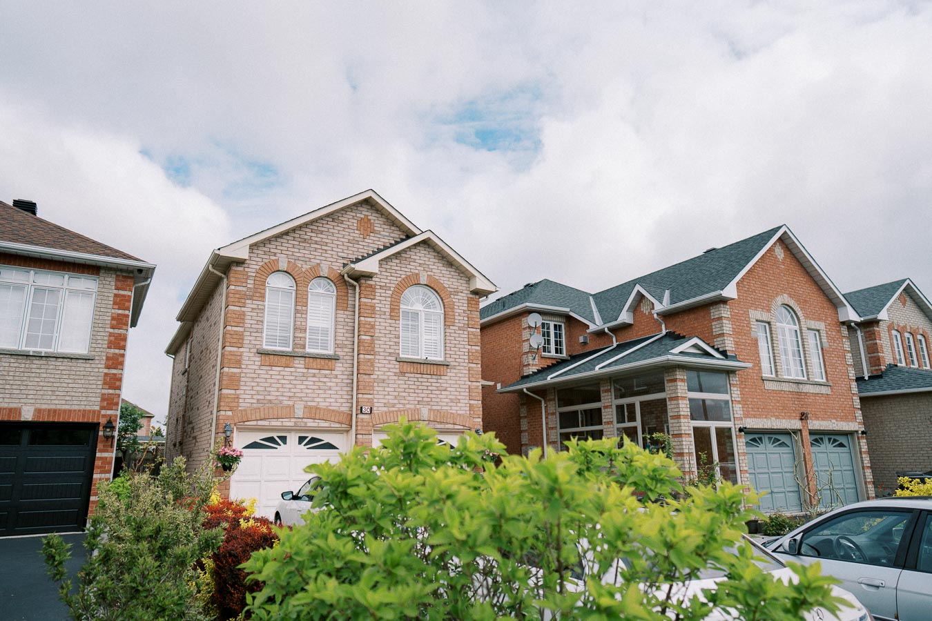 Suburban neighborhood with large brick houses and manicured shrubs under a partly cloudy sky.