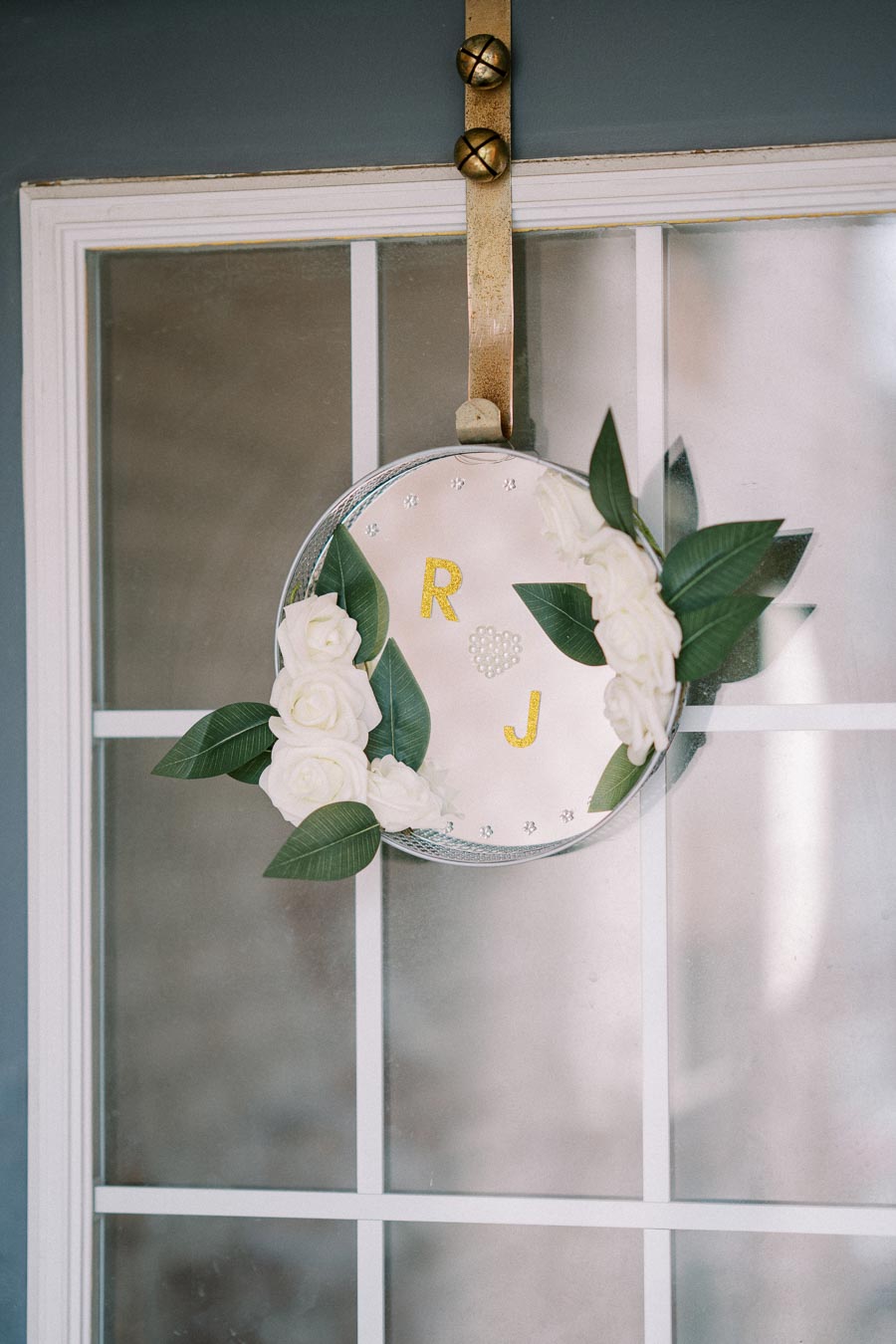 Wreath with white roses and green leaves hanging on a door, featuring the letters 'R' and 'J' in yellow against a round mirror background.