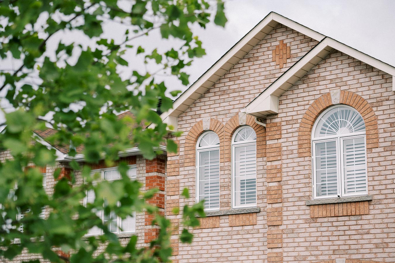 Close-up of a brick house facade with arched windows, partially obscured by green tree leaves in the foreground, under a cloudy sky.