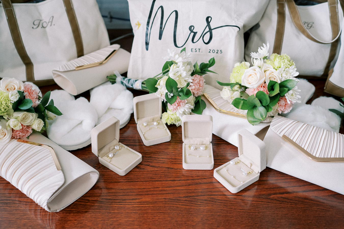 Bridal accessories and gifts arrangement featuring pearl jewelry in open boxes, white handbags, and floral bouquets with roses and greenery, set on a polished wooden table.