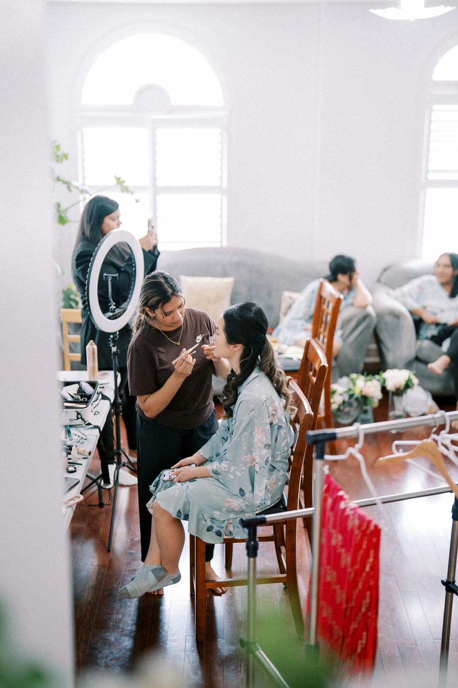 A makeup artist applying cosmetics to a woman in a floral robe, preparing for a special event, with a ring light and makeup tools in a bright room.