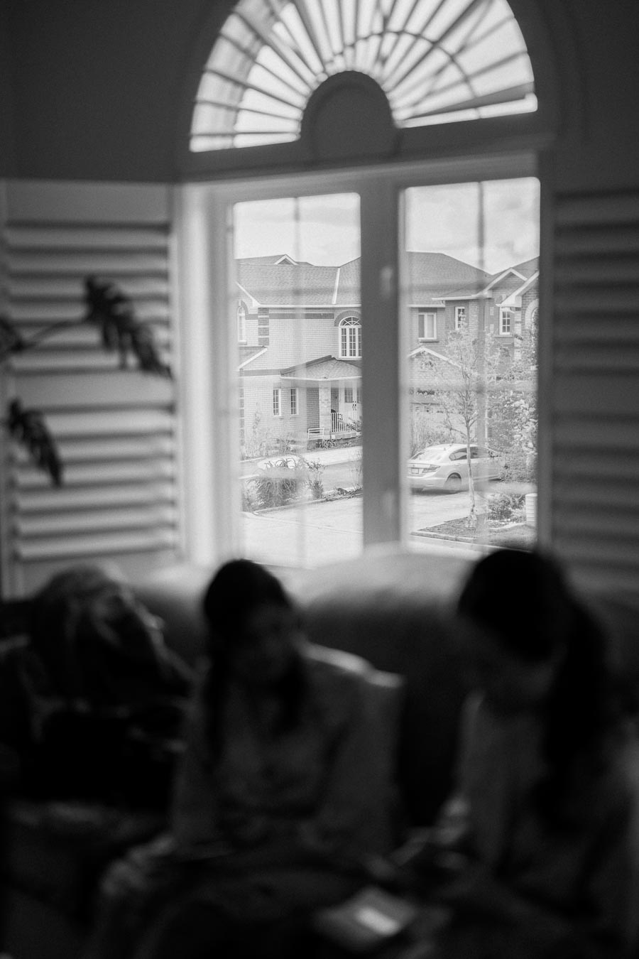 A black and white image showing the interior view of a room with two blurred figures sitting, looking out a large window that features an arched top, offering a view of suburban houses and a car parked outside.