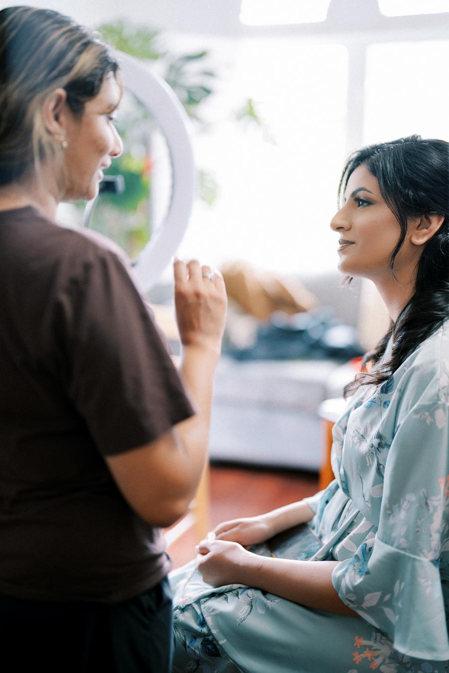A makeup artist applying cosmetics to a woman wearing a floral robe, preparing for an event in a brightly lit room.