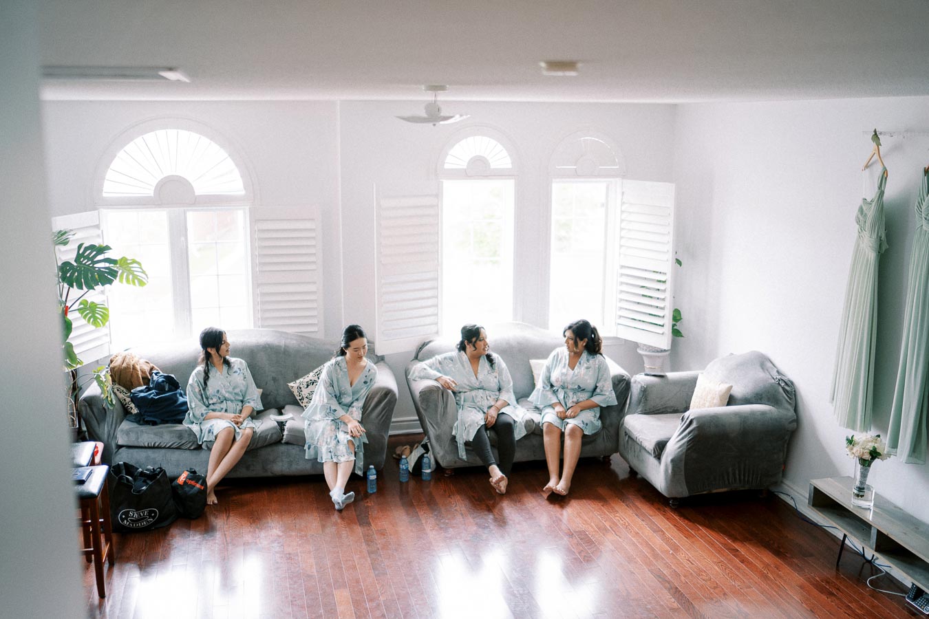 Four women in matching robes sit on couches in a well-lit living room, preparing for a wedding. Green bridesmaid dresses hang on the wall, and sunlight filters through window shutters, casting a warm glow on the wooden floor.