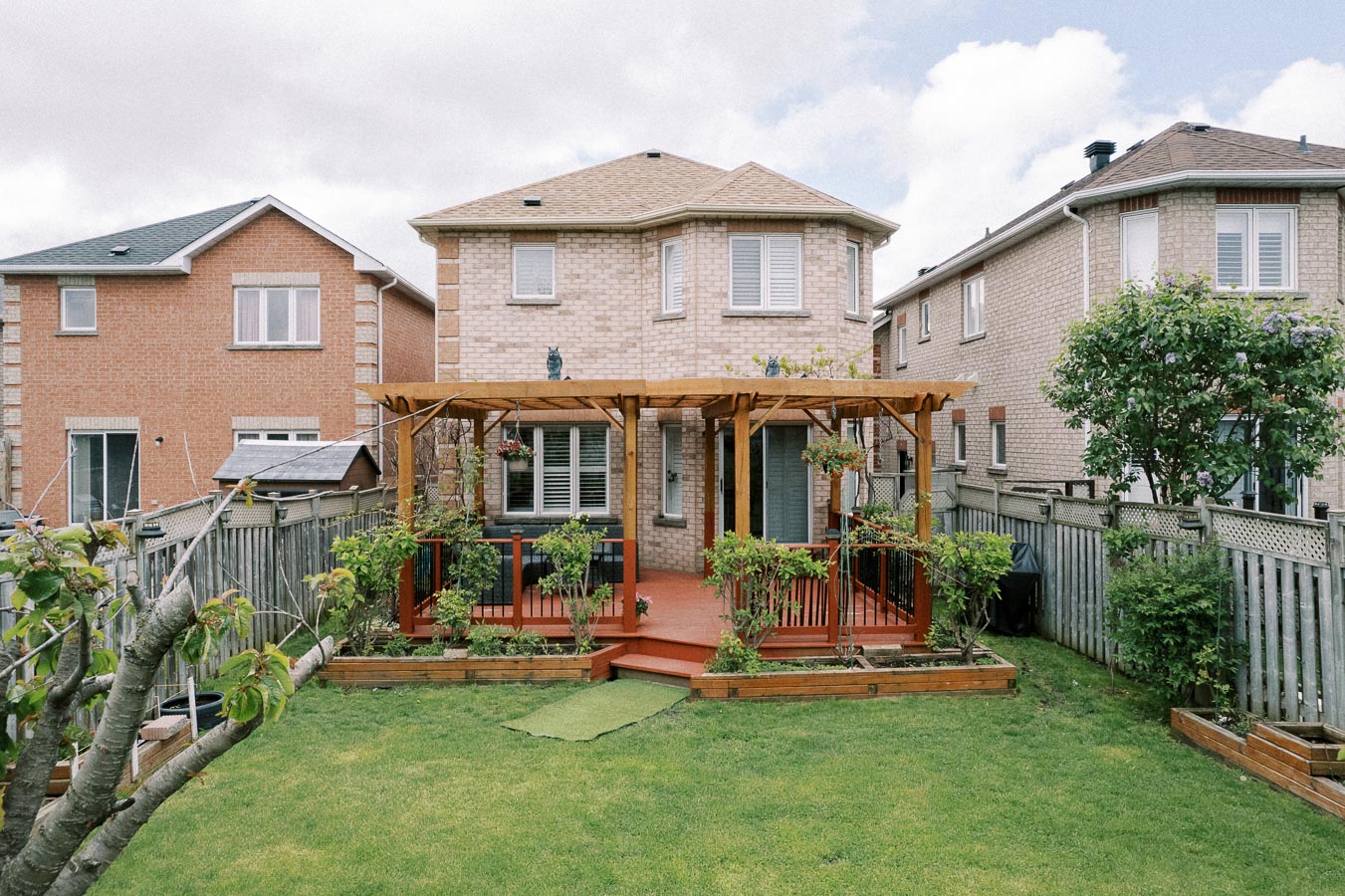 Two-story brick house with a wooden pergola on a well-maintained lawn, surrounded by a fence and neighboring homes, under a partly cloudy sky.