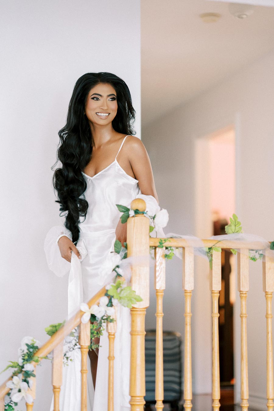 Bride in white silk robe with long black hair smiling on the decorated staircase, preparing for her wedding day indoors.