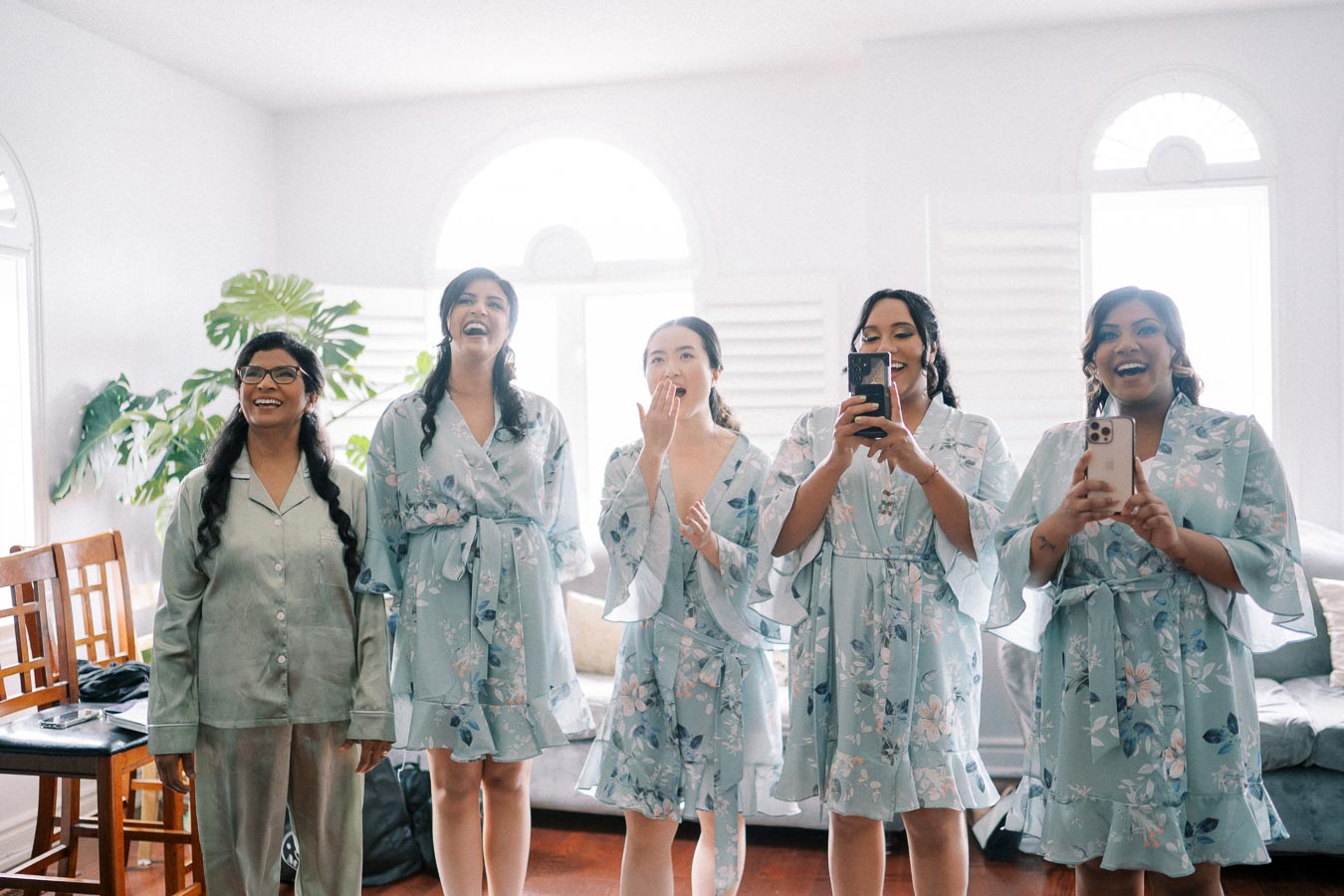 Five women in matching floral robes smile and capture the moment with smartphones in a bright room, featuring wooden chairs and lush green plants, creating a joyful ambiance.